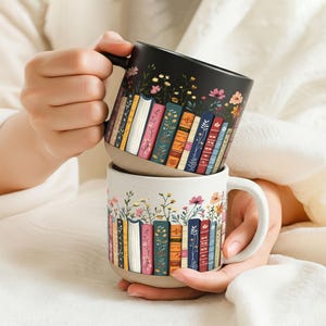 May include: Two ceramic mugs with a book and floral design. One mug is black with colorful books and flowers, the other is off-white with a similar design. Both mugs have a handle and are stacked.