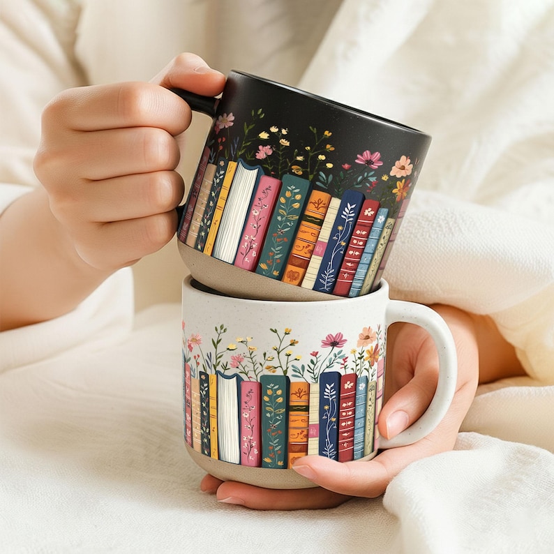 May include: Two ceramic mugs with a book and floral design. One mug is black with colorful books and flowers, the other is off-white with a similar design. Both mugs have a handle and are stacked.