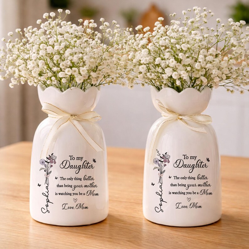 May include: Two white ceramic vases with a scalloped top, each tied with a cream ribbon and filled with white flowers. The vases have a floral design and text that reads "To my Daughter" and "Love Mom."