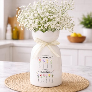K&ouml;nnte beinhalten: Wei&szlig;e Keramikvase mit gewelltem Rand, gebunden mit einem cremefarbenen Band, gef&uuml;llt mit wei&szlig;en Blumen. Die Vase ist mit den Worten "First Mom - Now Grandma" und floralen Illustrationen mit Namen verziert.