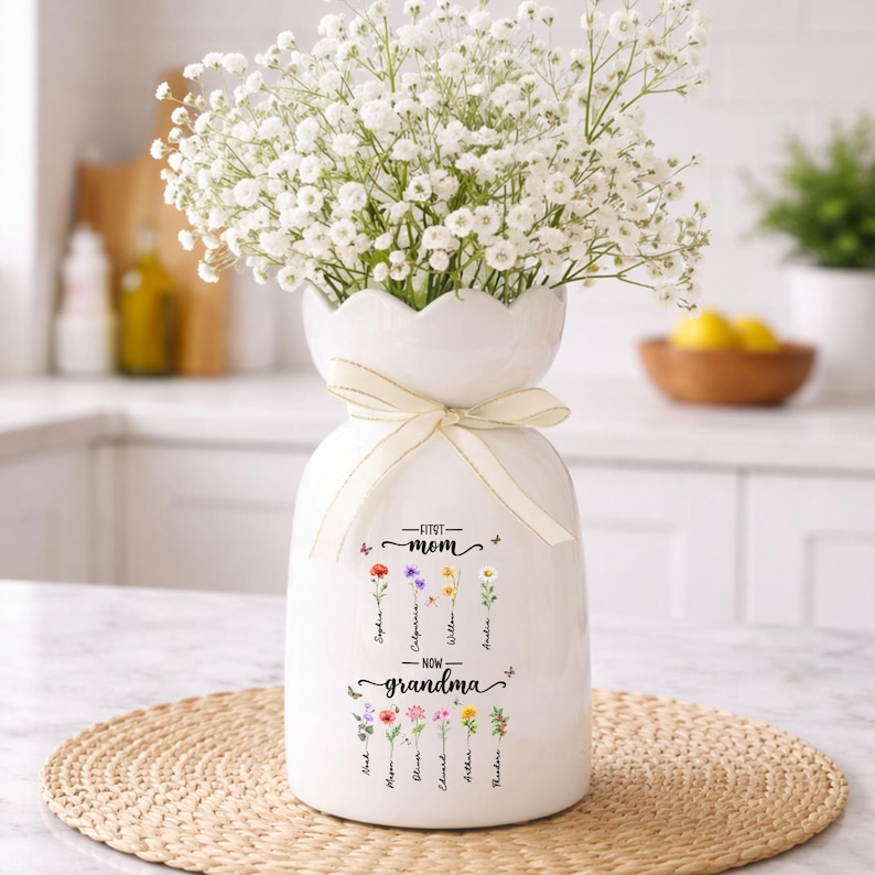 K&ouml;nnte beinhalten: Wei&szlig;e Keramikvase mit gewelltem Rand, gebunden mit einem cremefarbenen Band, gef&uuml;llt mit wei&szlig;en Blumen. Die Vase ist mit den Worten "First Mom - Now Grandma" und floralen Illustrationen mit Namen verziert.