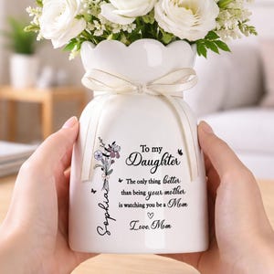 May include: White ceramic vase with a scalloped rim, tied with a cream-colored ribbon, and filled with white flowers. The vase has a floral design and the text "To my Daughter" with a personalized name and message.