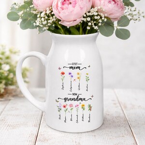 K&ouml;nnte beinhalten: Wei&szlig;er Keramikkrug mit Henkel, verziert mit dem Text "First Mom - Now Grandma" und floralen Illustrationen. Der Krug enth&auml;lt rosa Pfingstrosen und Gr&uuml;nzeug und bildet einen dekorativen Blumenstrau&szlig;.