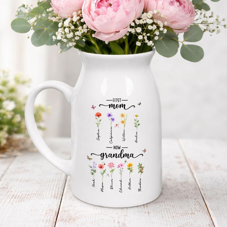 K&ouml;nnte beinhalten: Wei&szlig;er Keramikkrug mit Henkel, verziert mit dem Text "First Mom - Now Grandma" und floralen Illustrationen. Der Krug enth&auml;lt rosa Pfingstrosen und Gr&uuml;nzeug und bildet einen dekorativen Blumenstrau&szlig;.