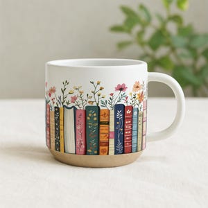 May include: White ceramic mug featuring a colorful design of books and flowers. The base of the mug is a natural tan color. The books are various colors with floral details. The handle is white.