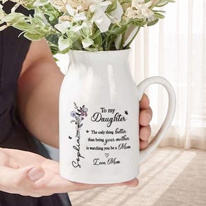 May include: A white ceramic vase with a handle, filled with white flowers. The vase is decorated with a floral design and the text "To my Daughter" along with a personalized message. The vase is held by a person.