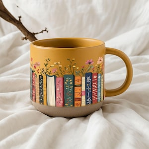 May include: Mustard yellow ceramic mug featuring a design of colorful books with flowers growing from the top. The mug has a tan base and a matching handle. The books have various floral and patterned designs.