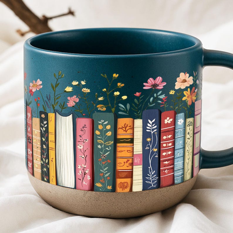 May include: A teal ceramic mug with a floral and book design. The mug features a row of colorful books with floral accents and a dark teal handle. The bottom of the mug is a light brown color.
