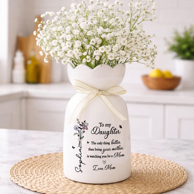 May include: White ceramic vase with a scalloped top, filled with white flowers. The vase has a cream-colored ribbon and a floral design with text that reads "To my Daughter" and "Love, Mom".