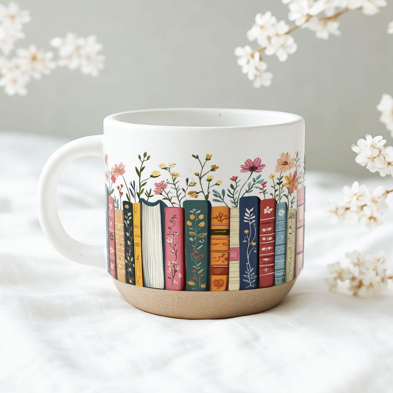 May include: White ceramic mug with a wraparound design of colorful books and floral accents. The books are in various colors, and the flowers include pink, yellow, and orange blooms. The mug has a tan base and a white handle.