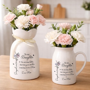 May include: Two white ceramic vases with floral arrangements. One vase is a unique shape with a ribbon, and the other is a mug shape. Both vases have text that reads "To my Daughter" and "Love Mom."