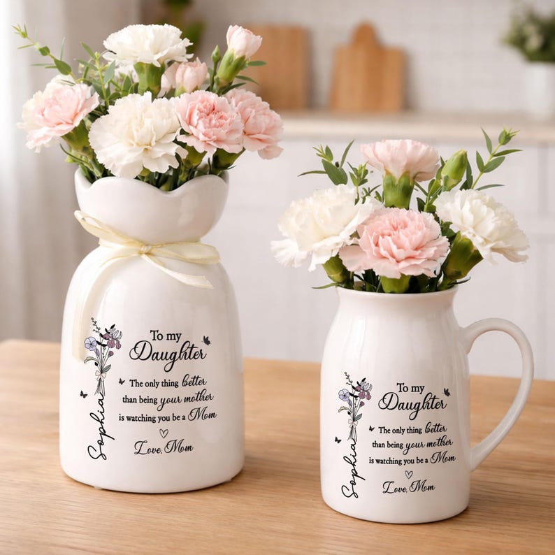 May include: Two white ceramic vases with floral arrangements. One vase is a unique shape with a ribbon, and the other is a mug shape. Both vases have text that reads "To my Daughter" and "Love Mom."