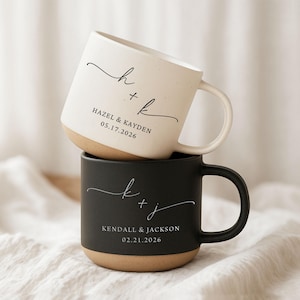 May include: Two stacked ceramic mugs. The top mug is off-white with black script reading "h + k" and "HAZEL & KAYDEN 05.17.2026". The bottom mug is black with "k + j" and "KENDALL & JACKSON 02.21.2026". Both have a tan base.