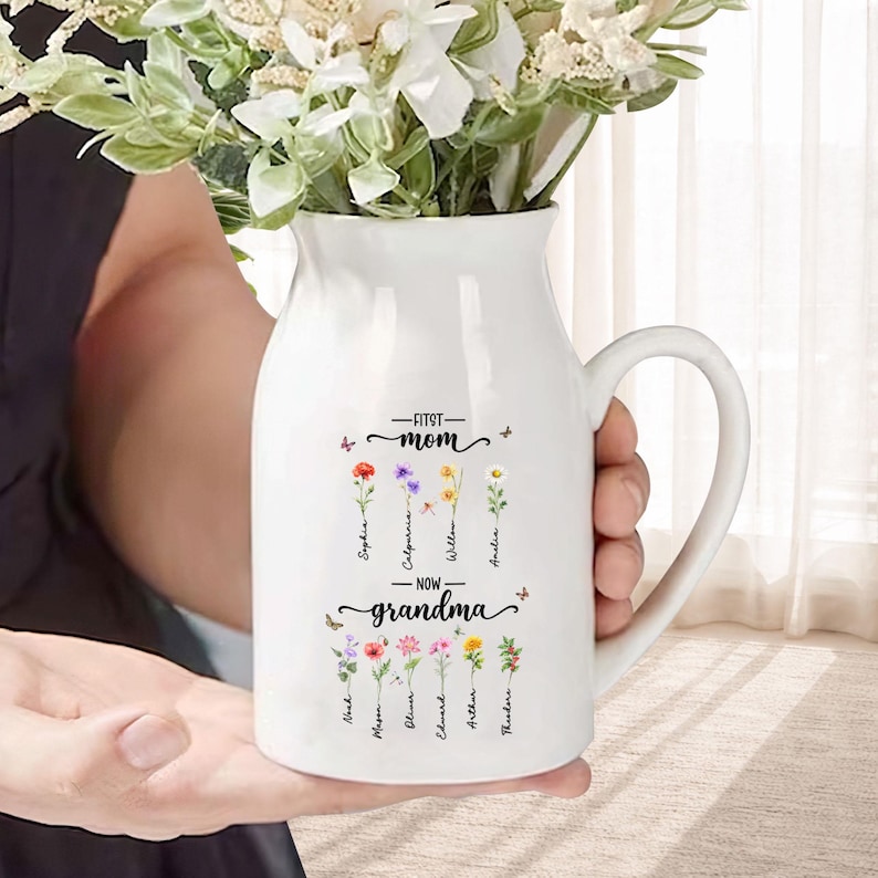 K&ouml;nnte beinhalten: Wei&szlig;er Keramikkrug mit Henkel, gef&uuml;llt mit wei&szlig;en Blumen. Der Krug ist mit den Worten "First Mom - Now Grandma" und floralen Illustrationen mit Namen verziert. Ein nachdenkliches Geschenk f&uuml;r eine Gro&szlig;mutter.