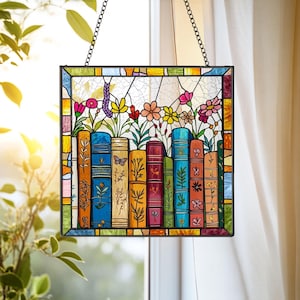 May include: Stained glass window panel featuring a colorful design of books with flowers. The books are various colors and have floral and butterfly details. The panel is framed with a black metal border and hangs from a chain.