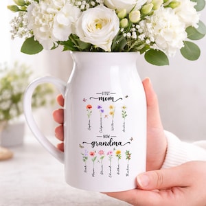 K&ouml;nnte beinhalten: Eine wei&szlig;e Keramikvase mit Henkel, gef&uuml;llt mit wei&szlig;en Blumen. Die Vase tr&auml;gt den Text "First Mom - Now Grandma" und Namen mit floralen Illustrationen. Die Vase wird von einer Person gehalten.