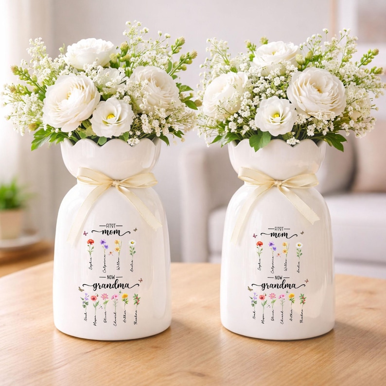 K&ouml;nnte beinhalten: Zwei wei&szlig;e Keramikvasen mit gewelltem Rand, jede mit einer beigen Schleife gebunden. Die Vasen sind mit wei&szlig;en Blumen und Gr&uuml;n gef&uuml;llt. Auf der Vase steht "First Mom - Now Grandma" mit Namen und floralen Illustrationen.
