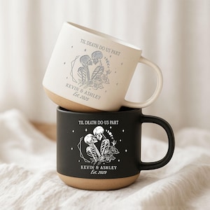 May include: Two stacked ceramic mugs. The top mug is off-white with a brown base, the bottom mug is black with a brown base. Both mugs feature a skeleton couple design with the text "TIL DEATH DO US PART" and "KEVIN & ASHLEY Est. 2025".