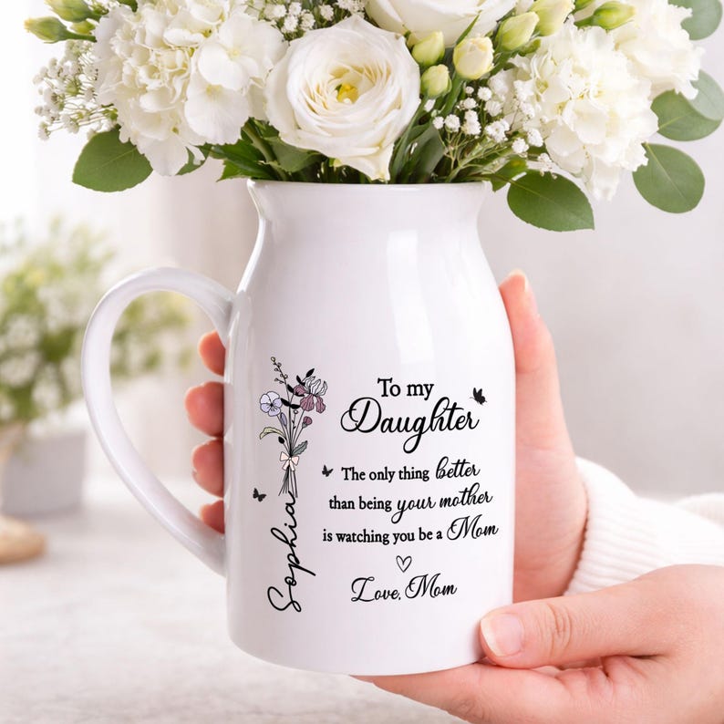 May include: White ceramic vase with a handle, filled with white flowers. The vase has a floral design and text that reads "To my Daughter" and "Love, Mom". A bouquet of flowers is also depicted.