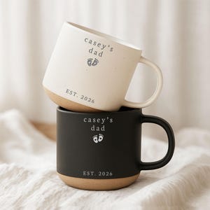 May include: Two ceramic mugs, one cream and one black, stacked on top of each other. The cream mug has a white handle and the black mug has a black handle. Both mugs have the text "casey's dad", baby footprints, and "EST. 2026".