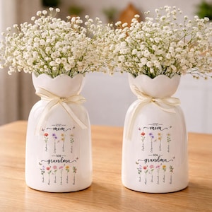 K&ouml;nnte beinhalten: Zwei wei&szlig;e Keramikvasen mit floralem Design und dem Text "First Mom - Now Grandma". Jede Vase ist mit wei&szlig;en Schleierkrautbl&uuml;ten gef&uuml;llt und mit einem cremefarbenen Band gebunden. Die Vasen stehen auf einer Holzoberfl&auml;che.