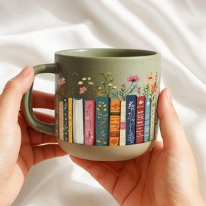 May include: A green ceramic mug featuring a design of colorful books with floral accents. The mug has a rounded shape with a handle and a light brown base. The books are decorated with various patterns and colors, and the flowers are in shades of pink, yellow, and white.