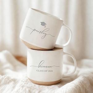 May include: Two white ceramic mugs stacked on top of each other. The top mug has the name "paisley" in cursive with a graduation cap graphic. The bottom mug has the name "brianna" in cursive with "CLASS OF 2026" below it. Both mugs have a tan base.