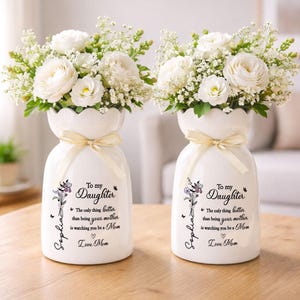 May include: Two white ceramic vases, each holding a bouquet of white flowers and greenery. The vases have a scalloped rim, a cream-colored ribbon, and a printed message: "To my Daughter" with a personalized name and a loving message.
