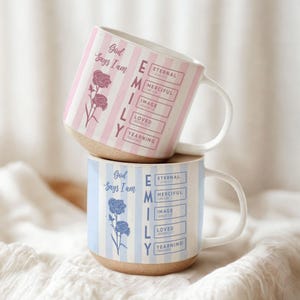 May include: Two ceramic mugs stacked on top of each other. The top mug has pink and white stripes with the text "God Says I Am" and "EMILY" with associated words. The bottom mug has blue and white stripes with similar text and a floral design.
