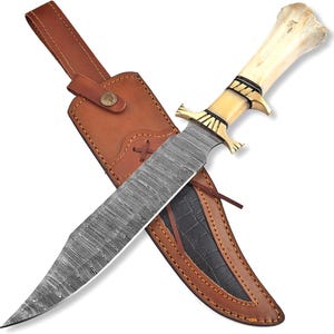 May include: A large, ornate knife with a patterned blade and bone handle, resting in a brown leather sheath. The handle features gold-colored accents. The sheath has a dark, textured interior and a leather strap.