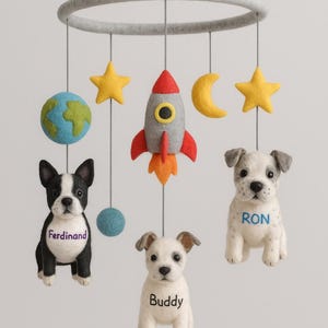 May include: A handmade baby mobile featuring felted dogs, a rocket ship, stars, a moon, and a globe. The dogs are named Ferdinand, Buddy, and Ron. The mobile is suspended from a gray felt ring.