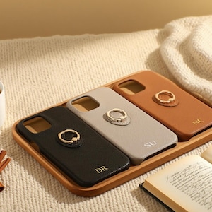 Könnte beinhalten: Drei personalisierte Handyhüllen in Schwarz, Grau und Braun, jede mit einem goldenen Ringhalter. Die Hüllen sind auf einem Holztablett angeordnet, wobei auf jeder Initialen eingraviert sind. Eine Tasse Kaffee, Zimtstangen und ein Buch sind ebenfalls im Bild.