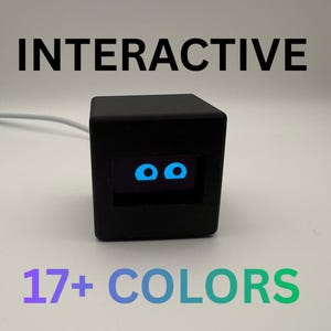May include: A black cube-shaped electronic device with a digital screen displaying two bright blue eyes. The text INTERACTIVE is printed above the device, and the text 17+ COLORS is printed below it. A white power cable extends from the side.