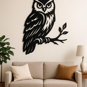 May include: A black metal owl wall art piece. The owl is perched on a branch with leaves. The artwork is mounted on a white wall above a beige sofa and a wooden coffee table. A lamp and a potted plant are also in the room.