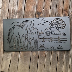 Può includere: Un pannello da parete rettangolare in metallo nero opaco con un design tagliato al laser raffigurante una giumenta e un puledro in un campo. La scena include una fattoria, una staccionata, un grande albero e uccelli, con fori di montaggio.