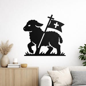 Op de afbeelding: Zwarte metalen wandkunst die een lam met een vlag met een kruis afbeeldt. Het lam loopt op gras. De vlag heeft een wit kruis op een zwarte achtergrond. Het kunstwerk is op een witte muur gemonteerd.