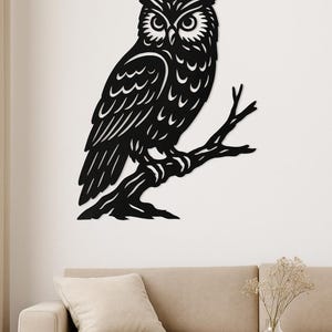 以下が含まれることがあります： 黒い金属製のフクロウの壁掛けアート。フクロウは枝に止まっており、羽と目の詳細なデザインが施されています。このアートワークは、ベージュのソファと木製のテーブルの上にある白い壁に取り付けられています。