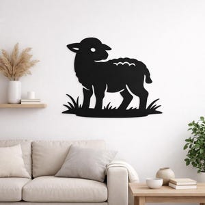 May include: A black metal lamb silhouette wall art piece. The lamb is standing on a patch of grass. The artwork is mounted on a white wall, with a sofa and decorative items in the background.