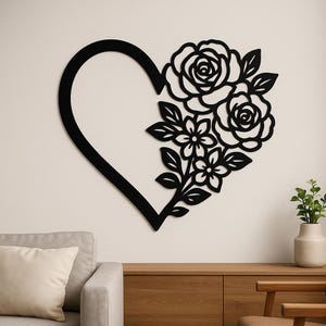 Puede incluir: Arte de pared en forma de corazón de metal negro con un diseño floral. El corazón está abierto por un lado y presenta rosas, flores pequeñas y hojas. La obra de arte está montada en una pared blanca sobre un mueble de madera y un sofá gris.