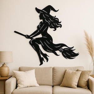 May include: Black metal wall art of a witch on a broomstick. The witch is wearing a pointed hat and a flowing dress. The artwork is mounted on a white wall above a beige sofa with cushions. A lamp and decorative plant are also visible.
