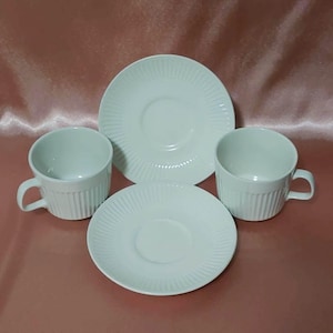 May include: A set of two white teacups with matching saucers. The cups and saucers have a ridged design. The set is displayed on a pink surface. The teacups have handles.
