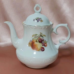 Puede incluir: Tetera de porcelana blanca con tapa, con un diseño de frutas de manzanas, cerezas y uvas. La tetera tiene un pico y un asa curvos, con ribete dorado. La tapa tiene un asa de anillo y un pequeño racimo de uvas.