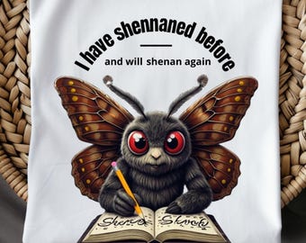 Puede incluir: Camiseta blanca con un diseño de mariposa de dibujos animados. La mariposa tiene grandes ojos rojos y sostiene un lápiz, escribiendo en un libro. El texto dice: "I have shennaned before and will shenan again".