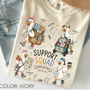 Può includere: Una t-shirt color avorio con una grafica di quattro anatre dei cartoni animati come squadra di supporto. Le anatre trasportano oggetti come uno zaino, un carretto e una tazza di caffè. Il testo include "SUPPORT SQUAD", "WE'VE GOT THIS!", "PLANNER", "ORGANIZER", "ENCOURAGER" e "COFFEE CAPTAIN" insieme a varie piccole icone.