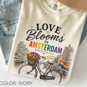 Może przedstawiać: T-shirt w kolorze kości słoniowej z grafiką zawierającą napis LOVE Blooms IN AMSTERDAM WORLDPRIDE 2026. Wzór przedstawia rower z koszem pełnym kolorowych tulipanów, tęczę i tęczową flagę na tle szkicu kamienic przy kanale.