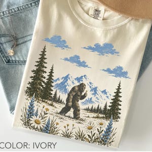 Bigfoot wildflower graphic t-shirt featuring a legendary Sasquatch walking through colorful mountain wildflowers, evergreen trees, and scenic alpine peaks in a vintage outdoor design on a unisex tee.
