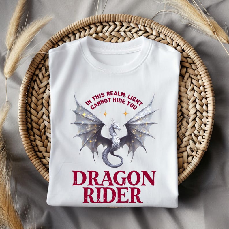 Puede incluir: Camiseta blanca con un drag&oacute;n plateado y el texto "IN THIS REALM, LIGHT CANNOT HIDE YOU". Debajo, el texto "DRAGON RIDER" en borgo&ntilde;a. La camiseta est&aacute; sobre una cesta tejida, con hierba seca.