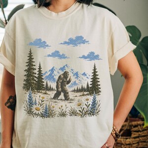 May include: A cream-colored short-sleeve t-shirt featuring a graphic print of a Bigfoot creature walking through a mountain landscape with pine trees, blue clouds, and a foreground filled with white daisies and blue lupine wildflowers.