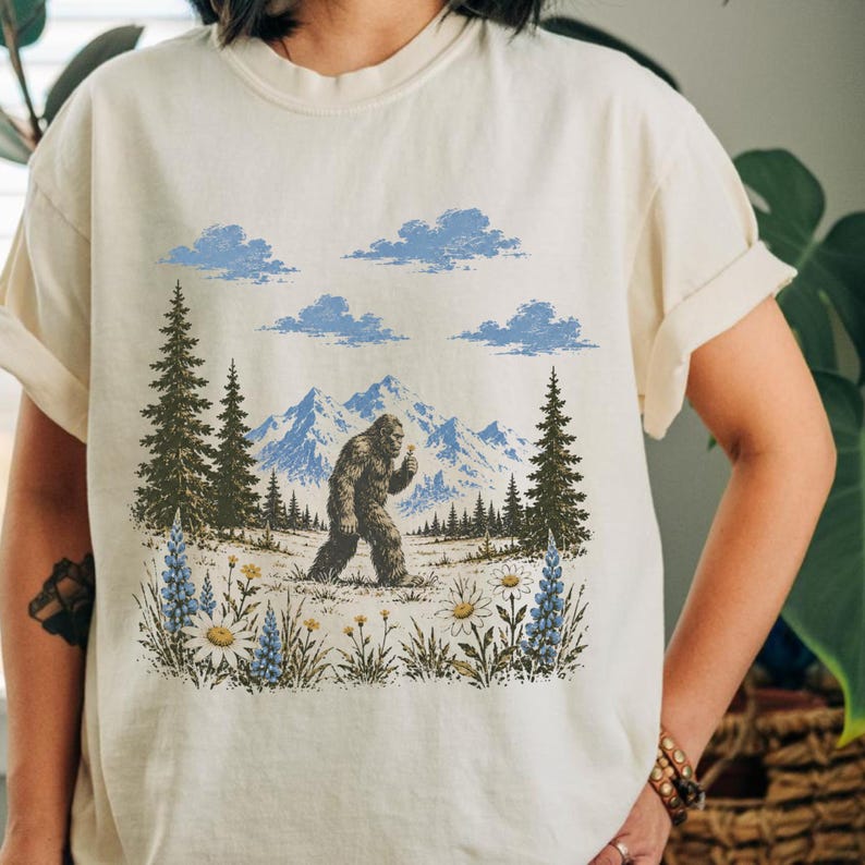 May include: A cream-colored short-sleeve t-shirt featuring a graphic print of a Bigfoot creature walking through a mountain landscape with pine trees, blue clouds, and a foreground filled with white daisies and blue lupine wildflowers.