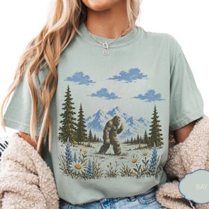 May include: A sage green t-shirt featuring a graphic print of a brown Bigfoot walking through a mountain landscape with pine trees, blue clouds, and wildflowers. The design includes a detailed illustration of a creature in a natural setting.